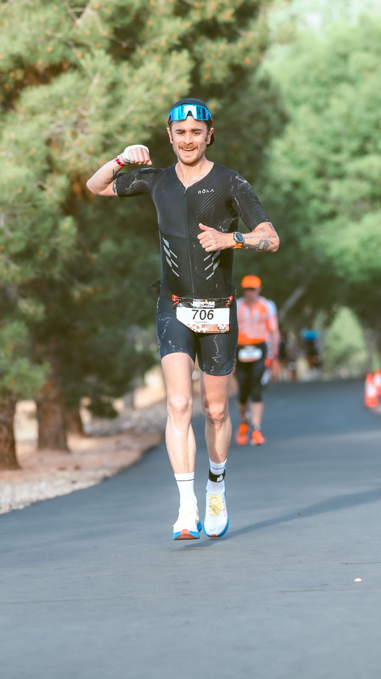 Smiling male triathlete wearing bib 706 flexes while running through a tree-lined course.