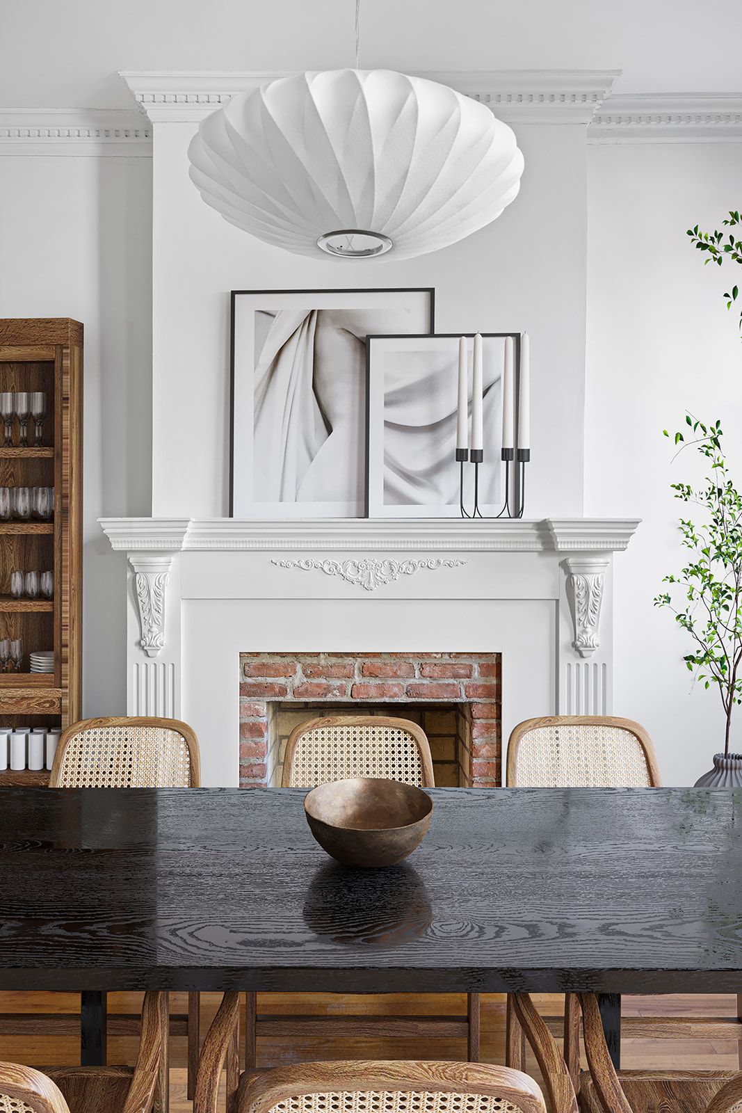 Elegant dining room with dark wood table, rattan chairs, white fireplace, brick interior, and abstract art above mantel.