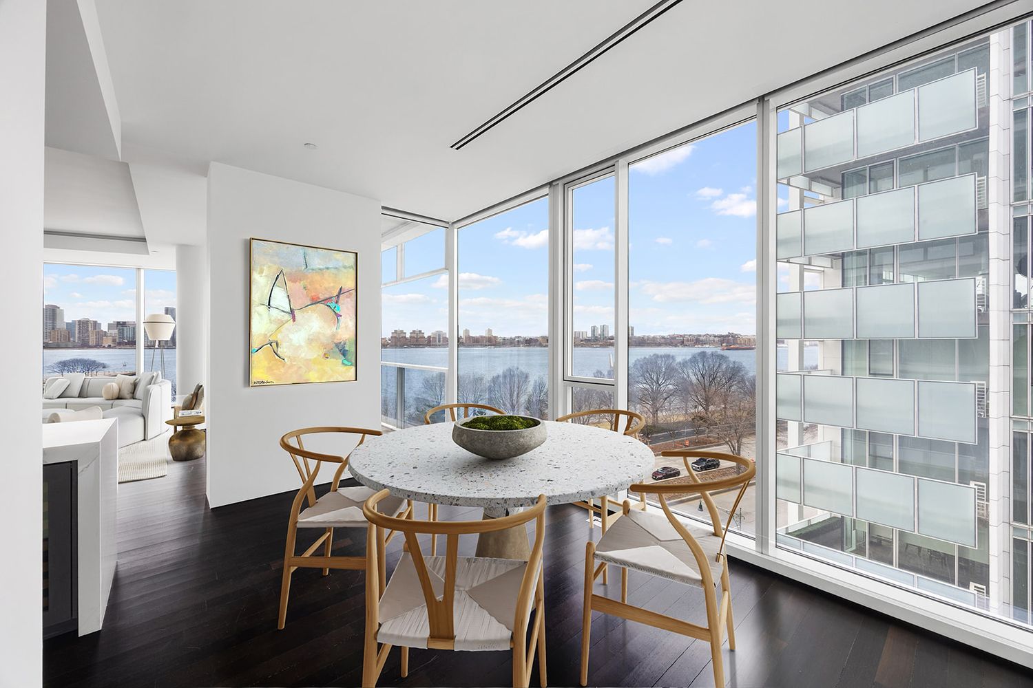 Modern dining area with floor-to-ceiling windows overlooking a river, featuring a round marble table and wooden chairs.