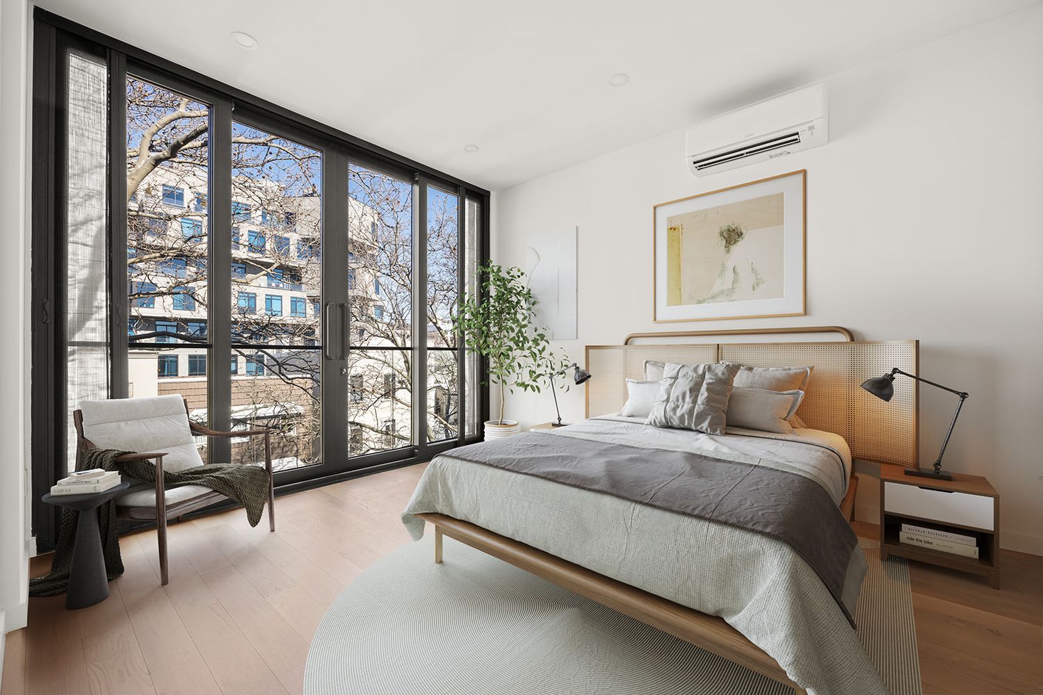 Modern bedroom with floor-to-ceiling windows, wood-framed bed, light hardwood floors, and a cozy reading chair with city views.