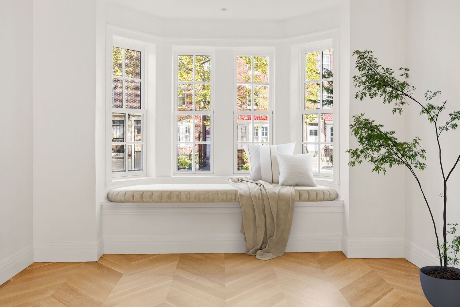 Bright bay window seat with cushion and throw blanket, herringbone wood floor, and indoor plant in minimalist white room.