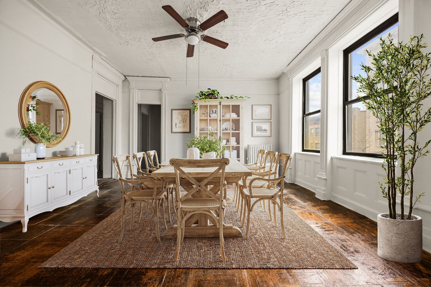 Bright dining room with hardwood floors, wainscoting, rattan chairs, oval rug, ceiling fan, and tall windows flooding natural light.