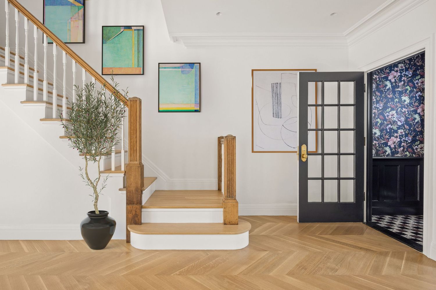 Bright entryway with herringbone wood floors, white staircase, modern art, olive tree, and bold floral wallpaper door.