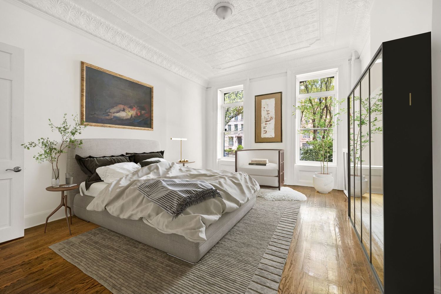 Elegant bedroom with gray upholstered bed, hardwood floors, ornate ceiling, large windows, and classic artwork in a bright space.