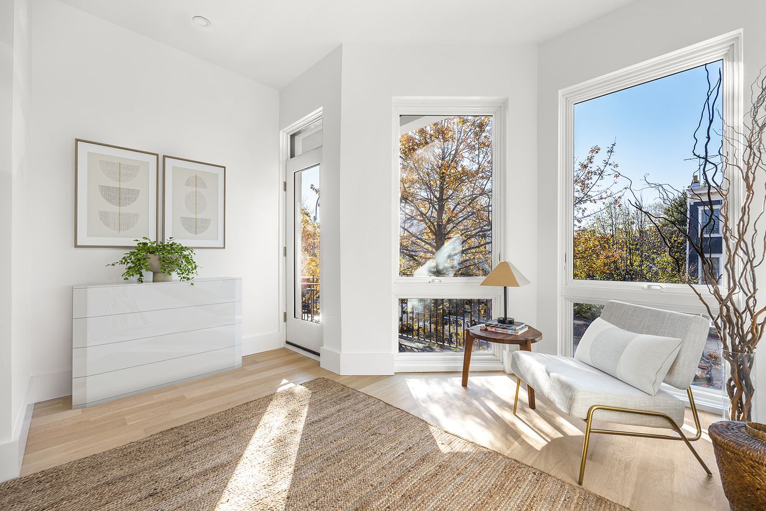 Bright modern bedroom with white walls, hardwood floors, jute rug, white dresser, and large windows with city views.