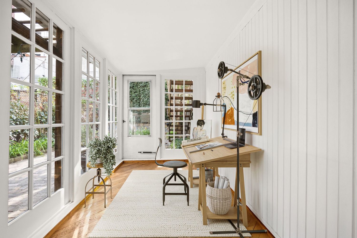 Bright home office with white shiplap walls, French doors, natural wood desk, industrial lamp, and white area rug.