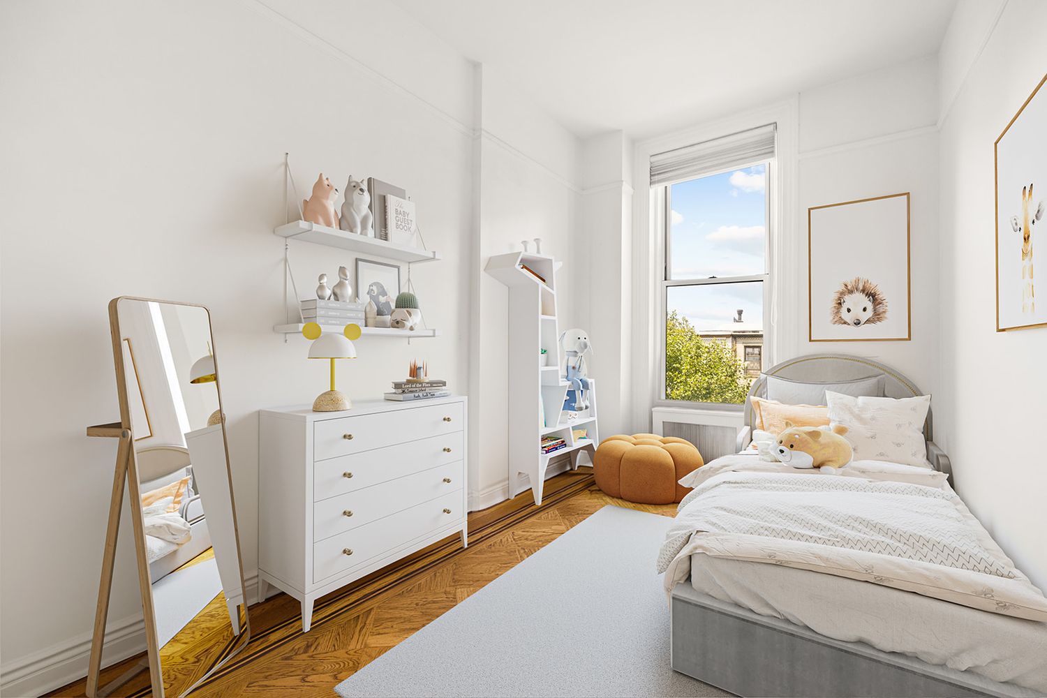 Bright child's bedroom with white dresser, leaning mirror, gray bed, parquet floors, and large window with natural light.