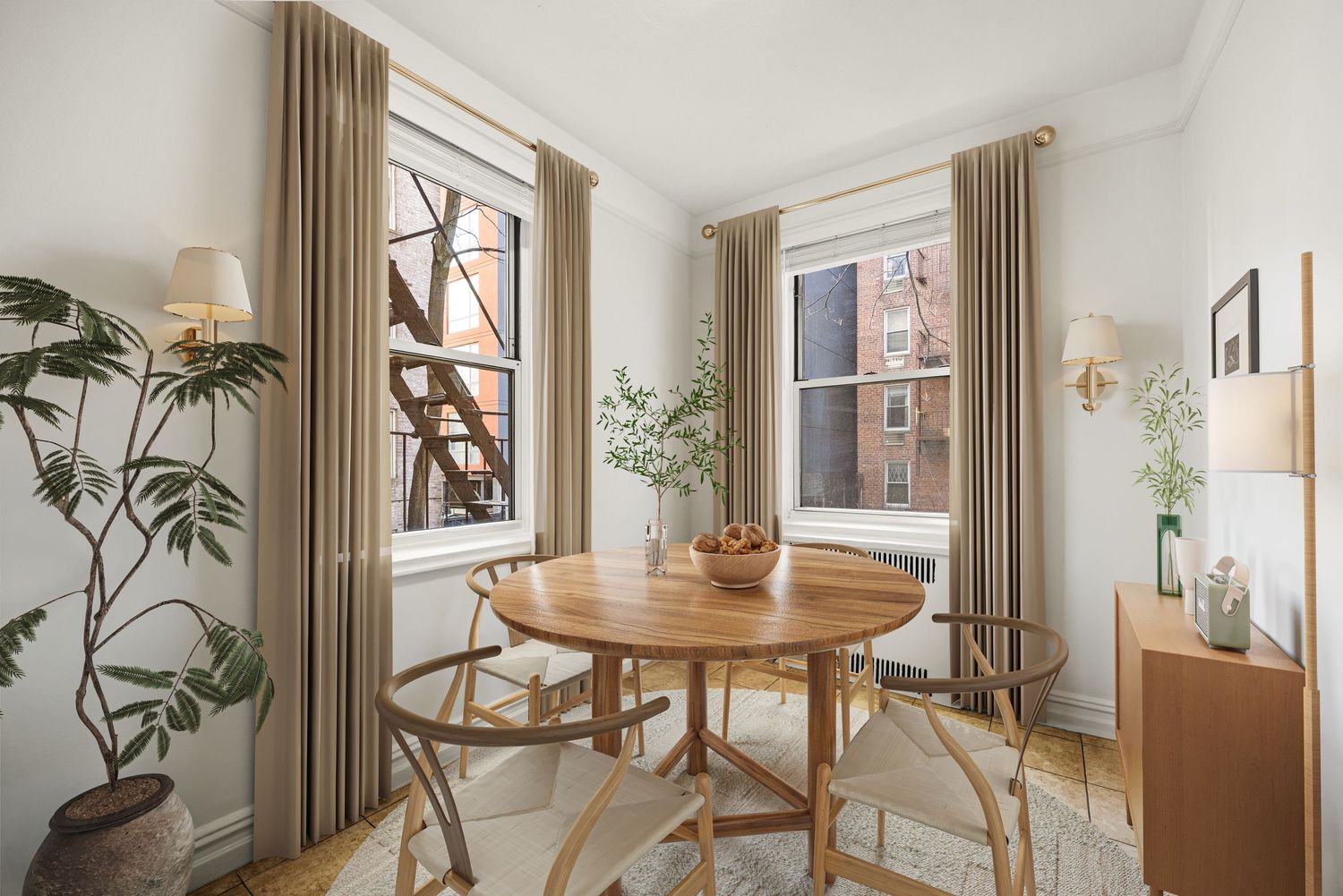 Bright dining nook with round wood table, wishbone chairs, tall windows with linen drapes, and a potted plant on herringbone floors.