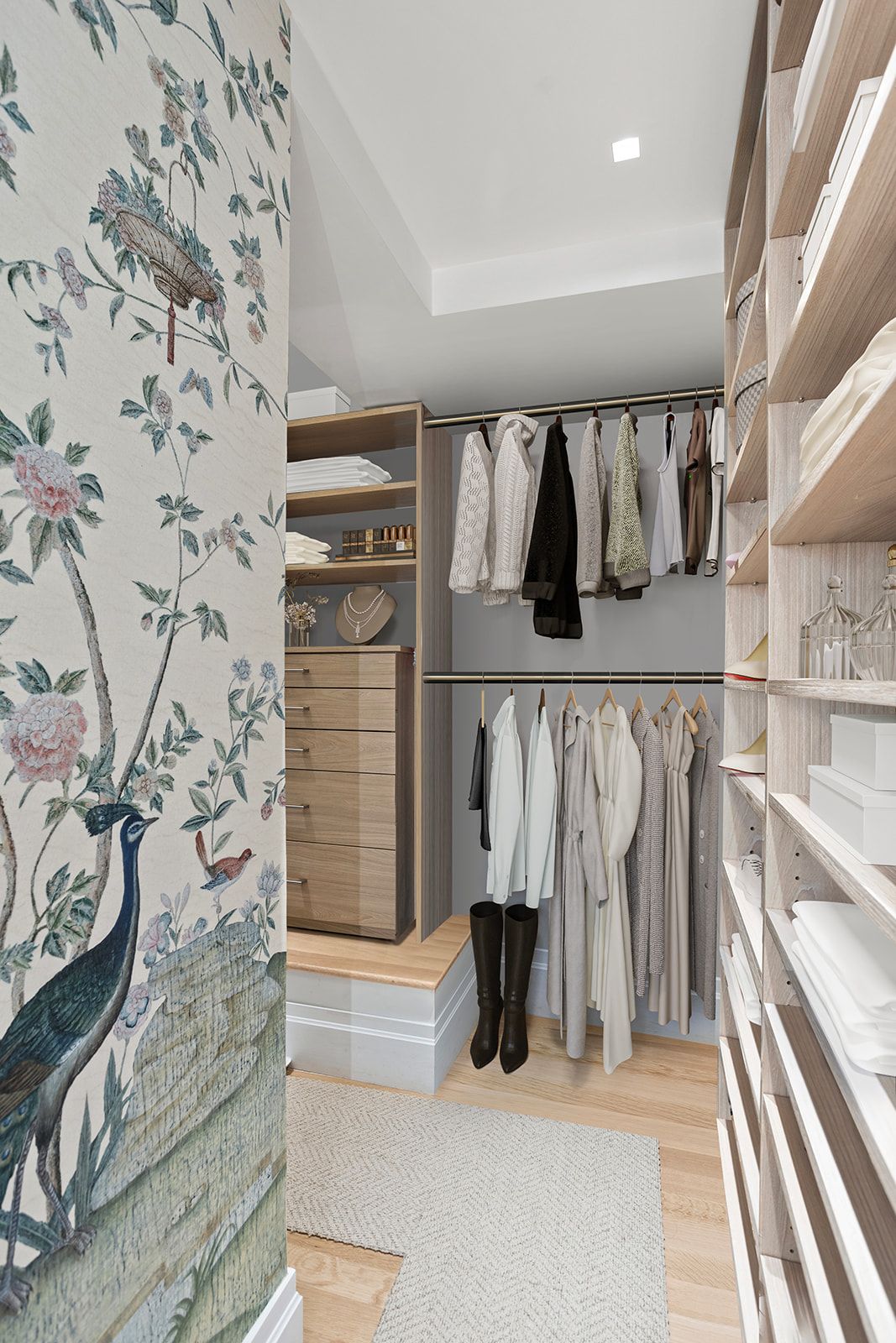 Elegant walk-in closet with floral wallpaper, light wood shelving, hanging clothes, and a small area rug on hardwood floors.
