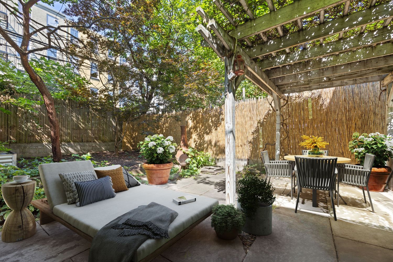 Serene outdoor patio with a daybed, dining set, lush greenery, and a pergola providing dappled shade.