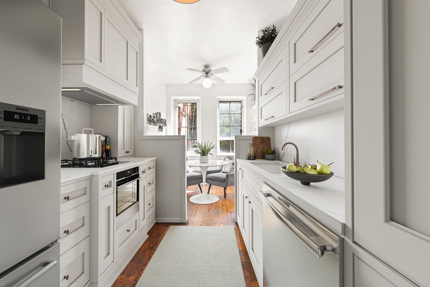 Bright galley kitchen with white shaker cabinets, stainless steel appliances, hardwood floors, and a cozy dining nook at the end.