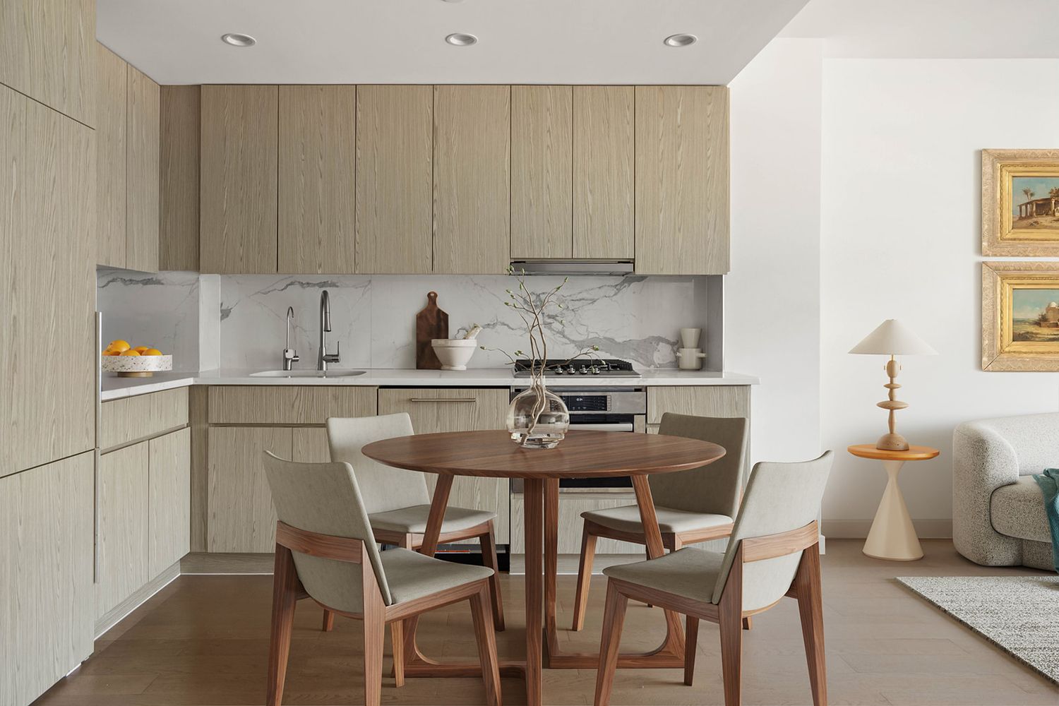 Modern open-plan kitchen and dining area with wood cabinetry, round walnut table, sage green chairs, and minimalist decor.