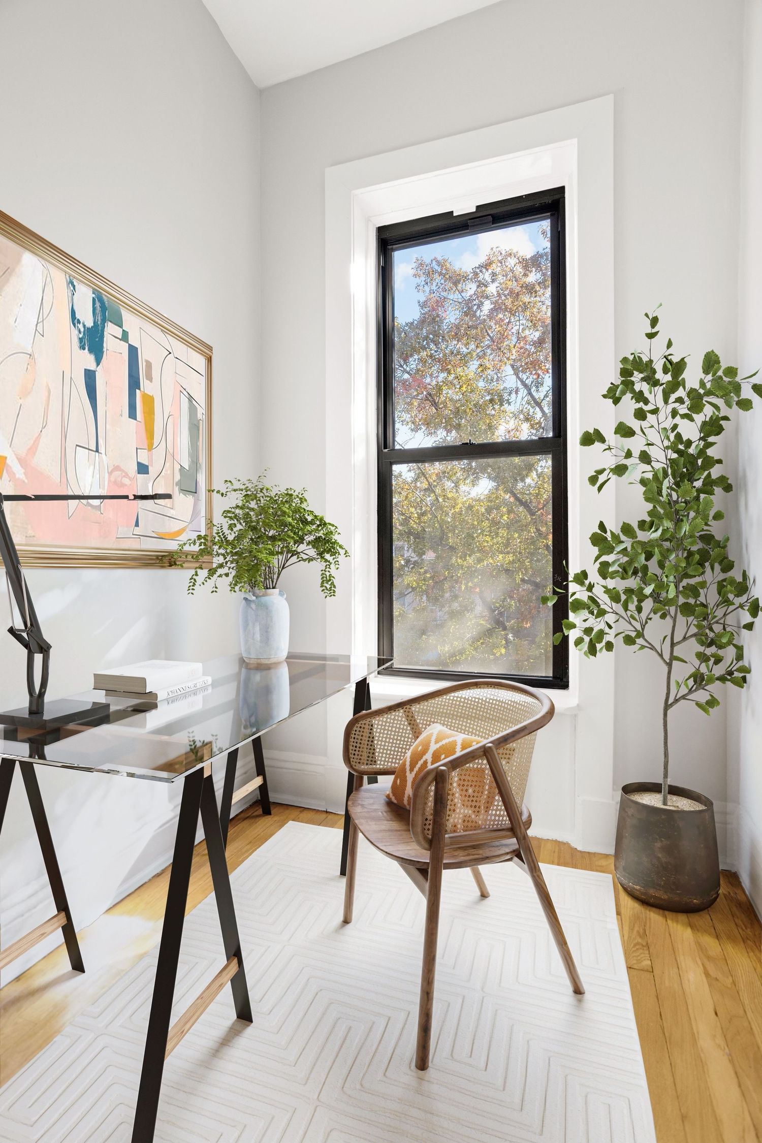 Bright home office with glass desk, wooden chair, abstract art, and tall window overlooking trees with hardwood floors.