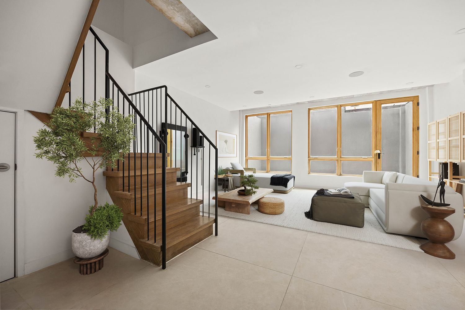 Modern open-plan living room with wood staircase, black railings, large windows, white sofa, and decorative bonsai tree.