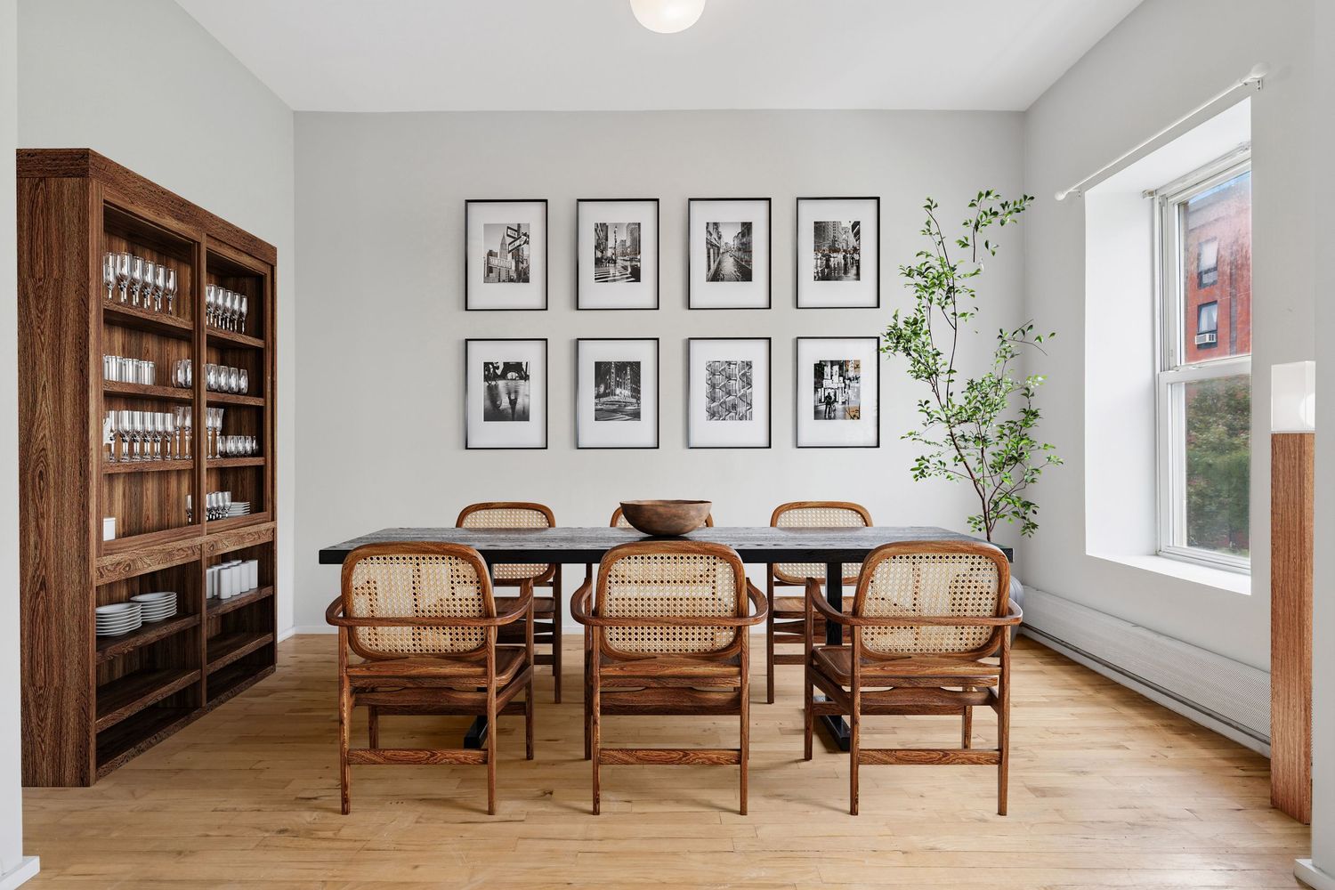 Modern dining room with wooden table, cane chairs, gallery wall, and hardwood floors beside a large window.