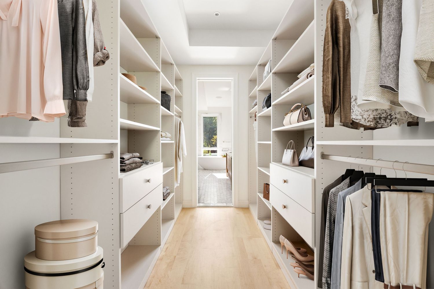 Bright walk-in closet with white built-in shelving, drawers, and hanging rods on both sides, featuring light hardwood floors.