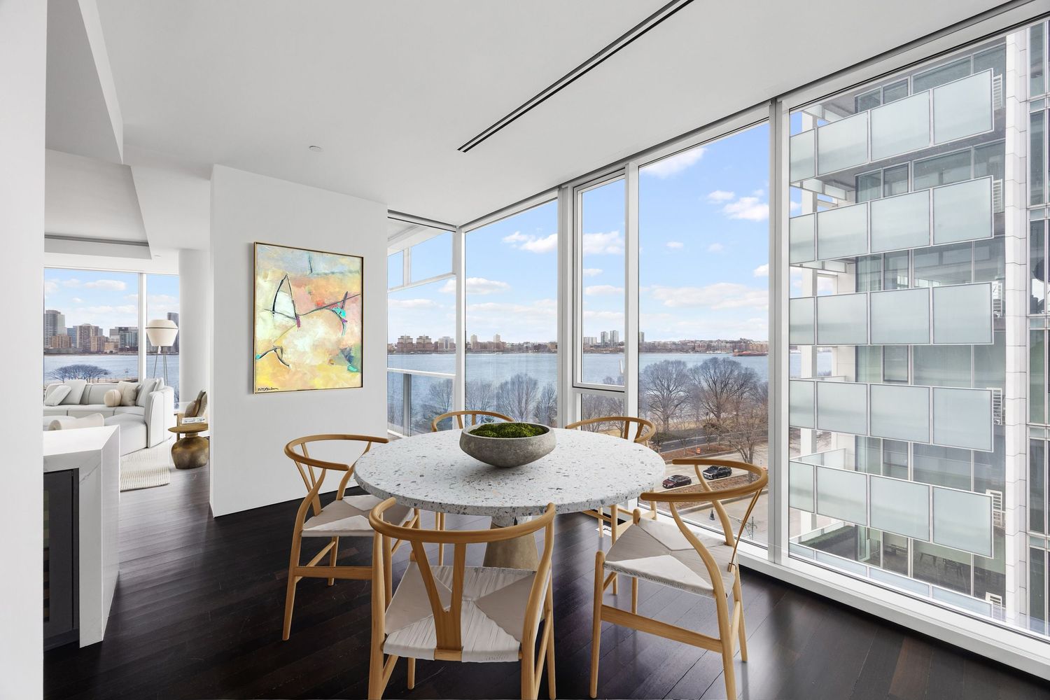 Modern dining area with floor-to-ceiling windows offering panoramic river views, featuring a marble table and wooden chairs.