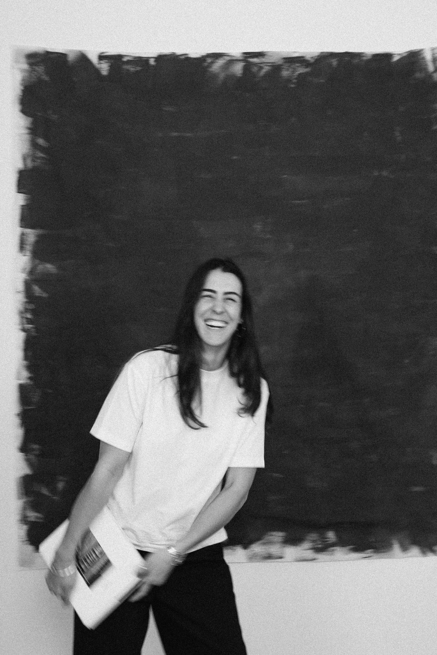 Black and white photo of a laughing woman in a white t-shirt holding a magazine before a dark abstract painting.