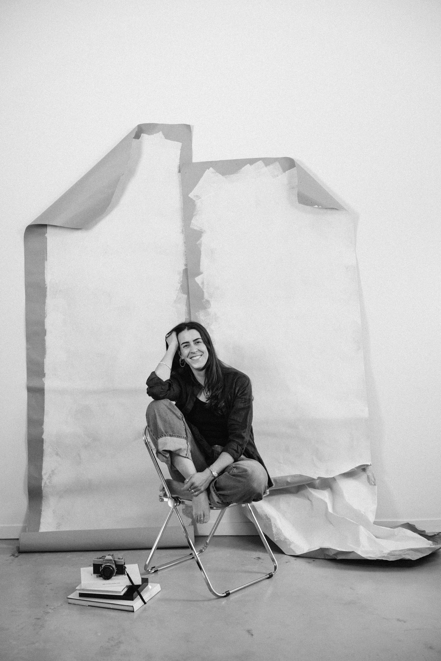 Smiling woman sitting on folding chair beside torn paper backdrop in black and white photo.