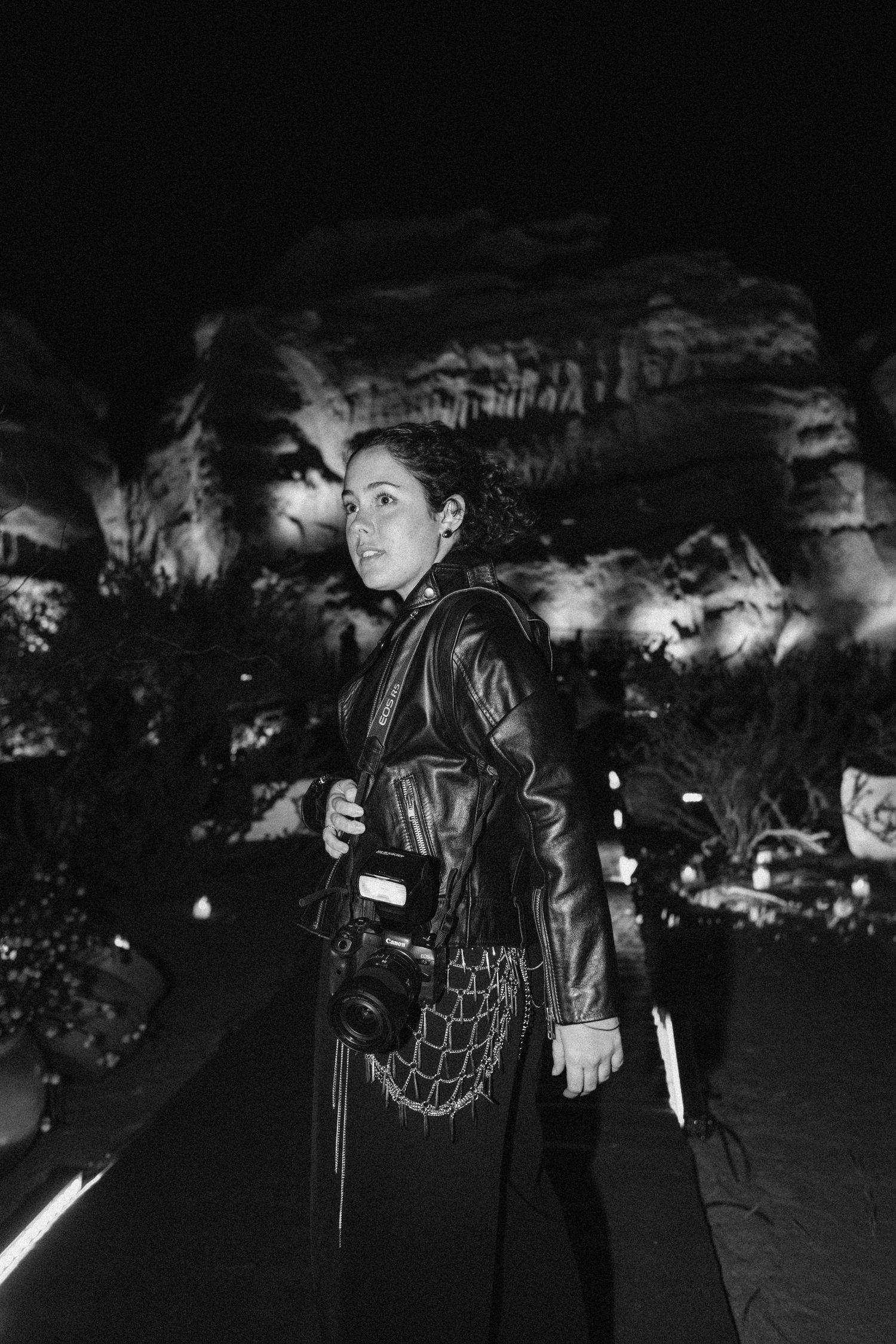 Woman with camera and mesh bag standing before illuminated rock formation at night event.