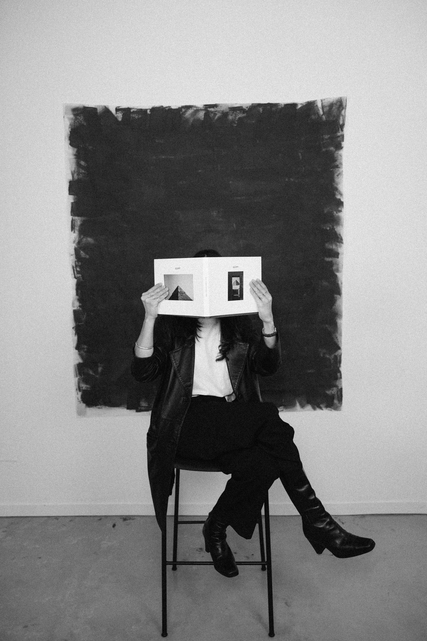 Person sitting on stool holding open book in front of face before dark painted wall.