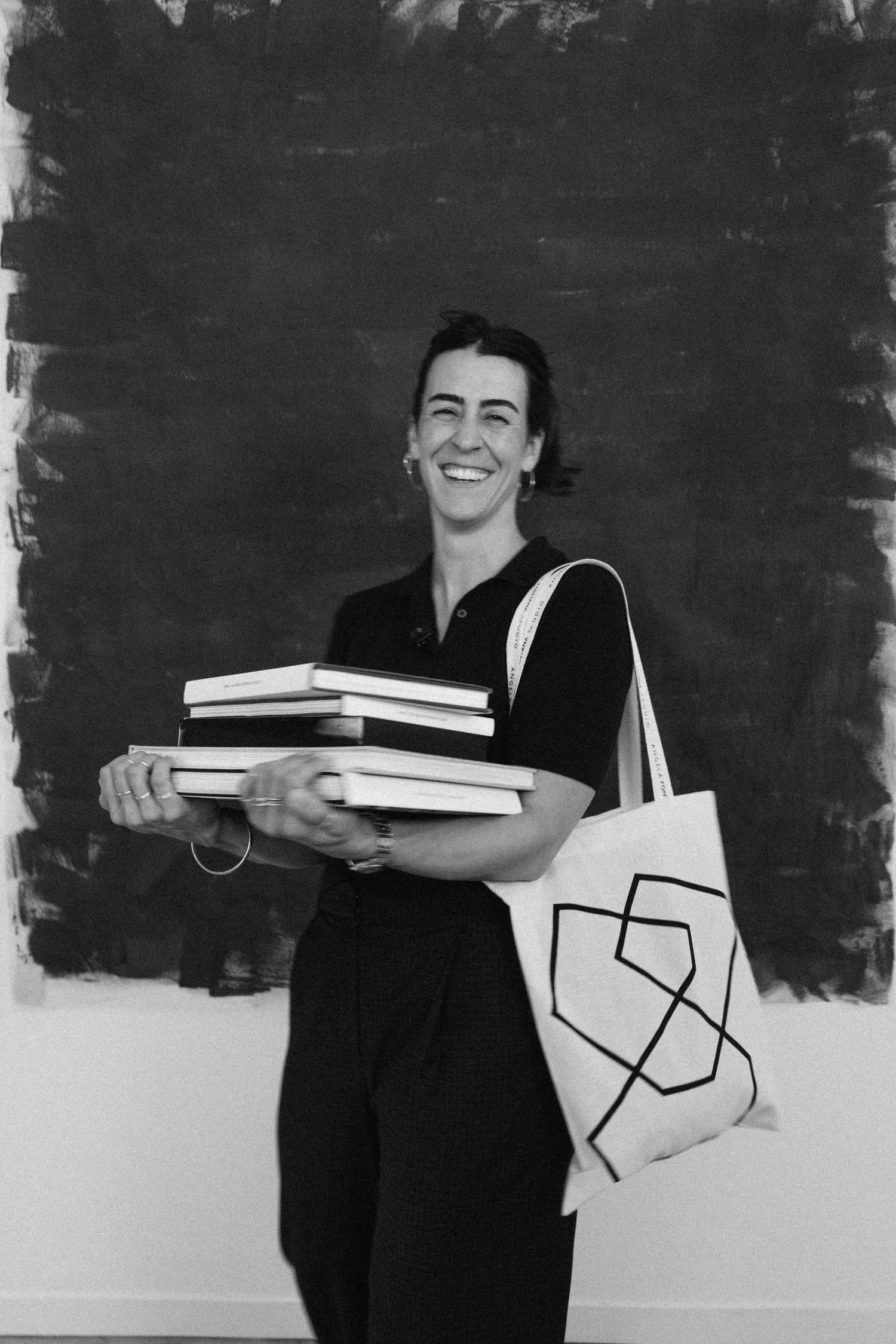 Laughing woman carrying books and geometric tote bag before dark painted canvas.