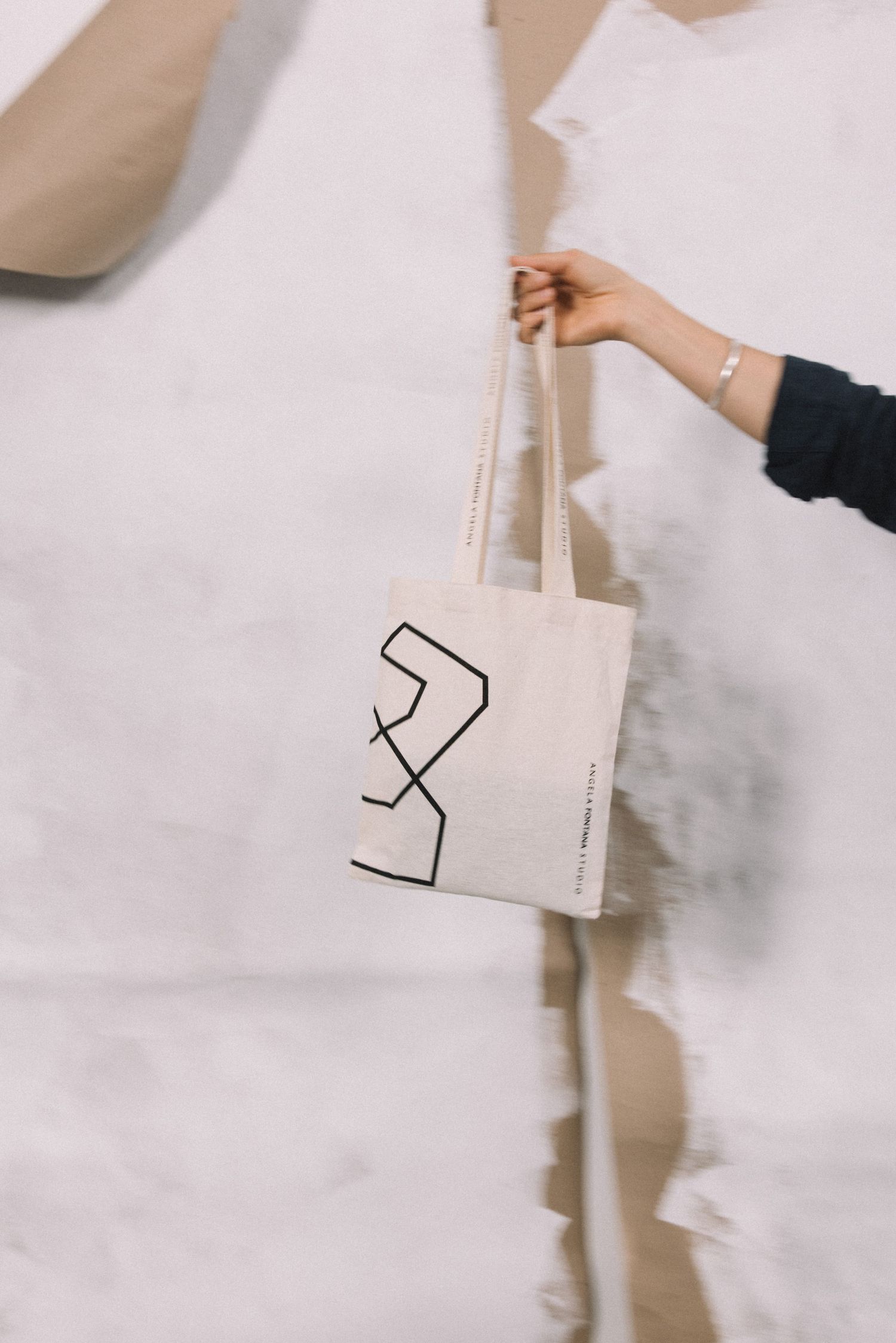 A hand holds a natural canvas tote bag with black geometric line art design against a torn paper backdrop.
