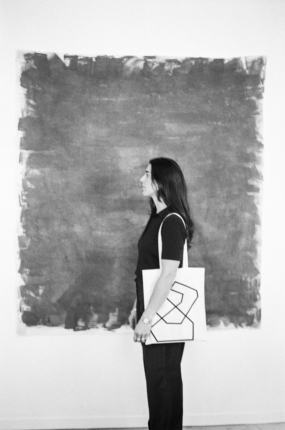 Woman in black holding geometric tote bag standing before large gray painted canvas.