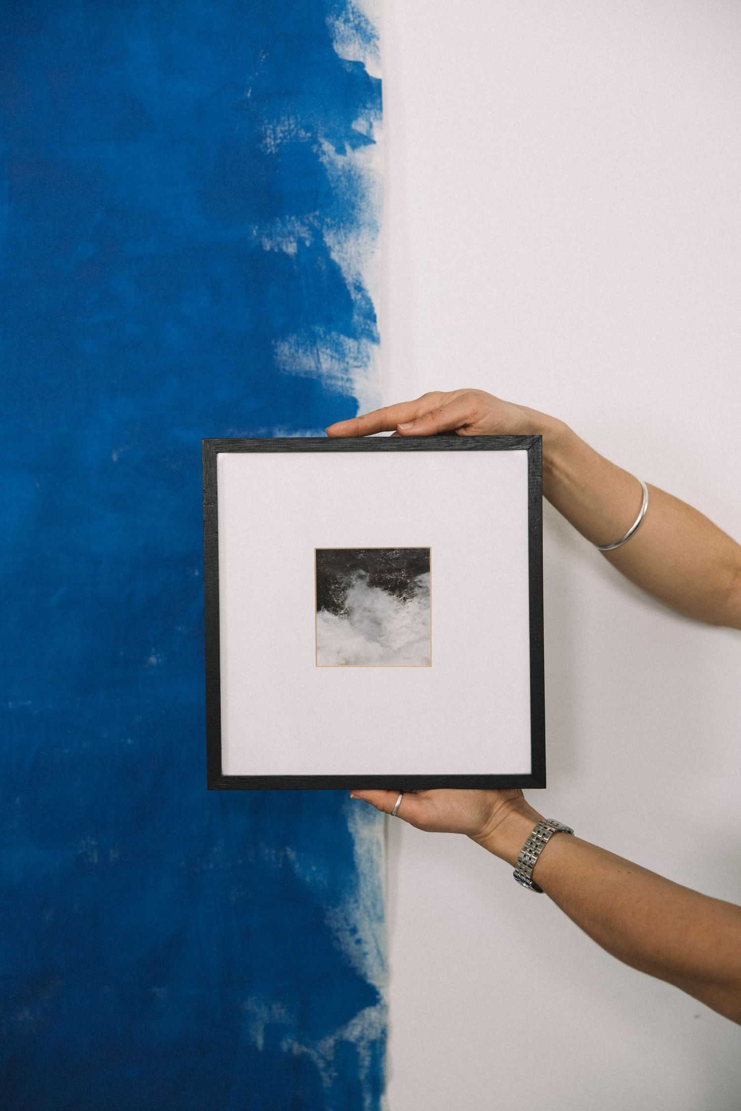 Two hands hold a black framed fine art print against a half blue painted white wall.