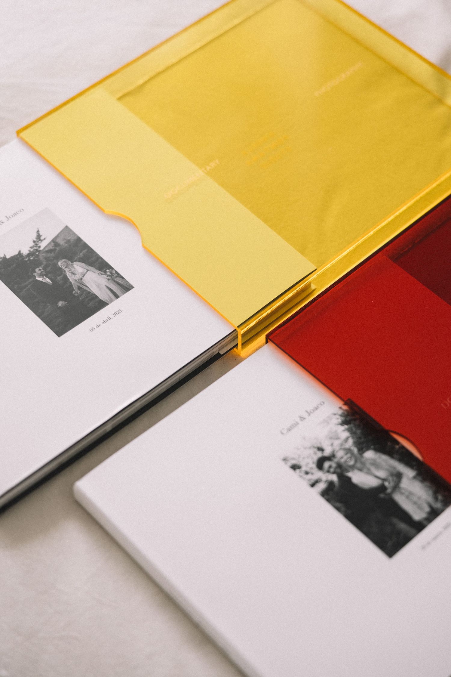 Three photo books with colorful transparent yellow and red acrylic covers lay overlapping on a white surface.