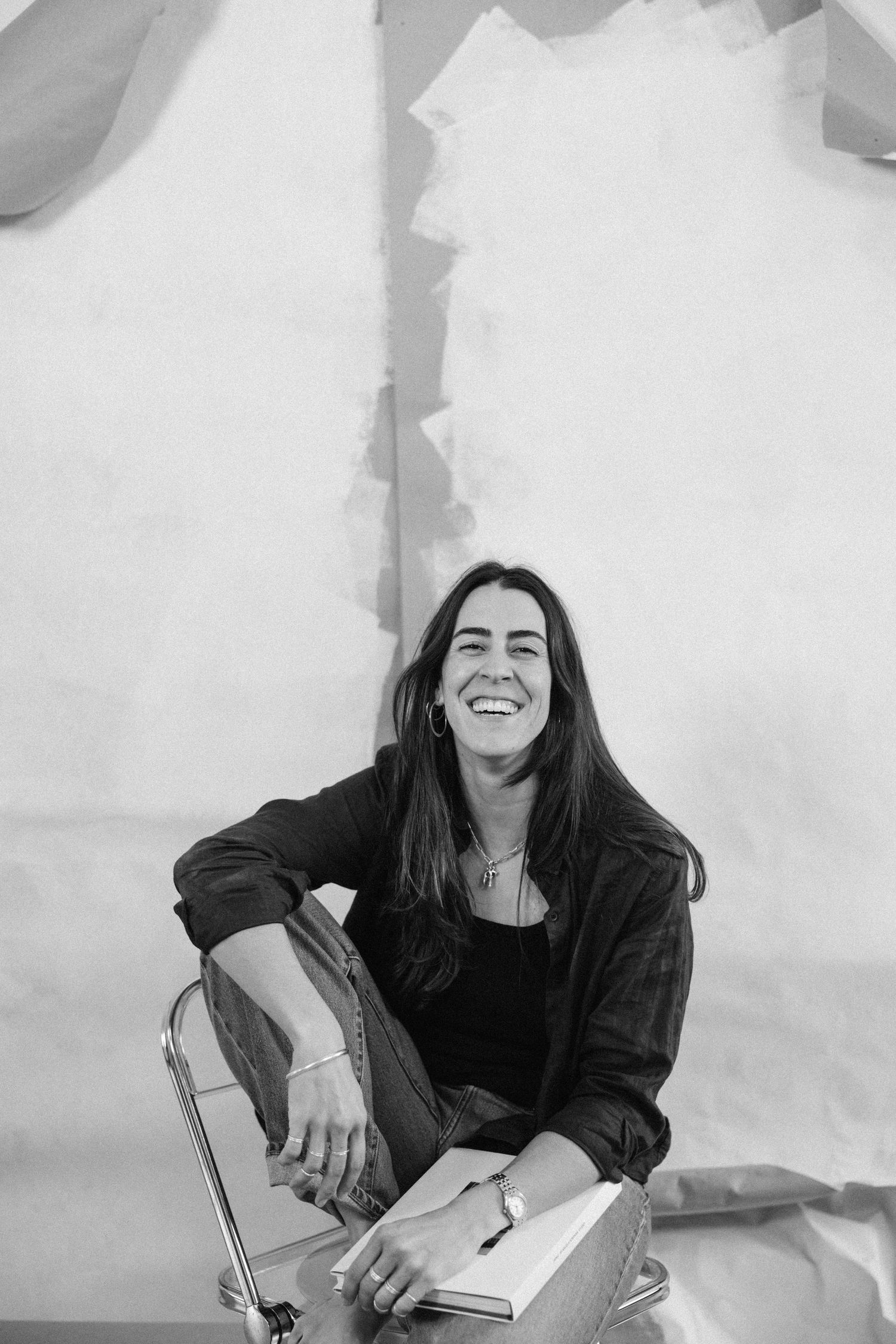 Black and white portrait of a smiling woman with long hair sitting cross-legged on a chair holding a notebook.