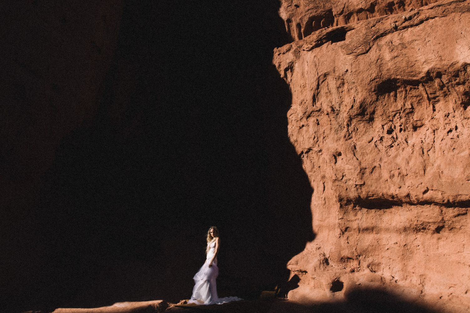 Bride in white wedding dress standing in sunlight between dramatic red sandstone canyon walls.
