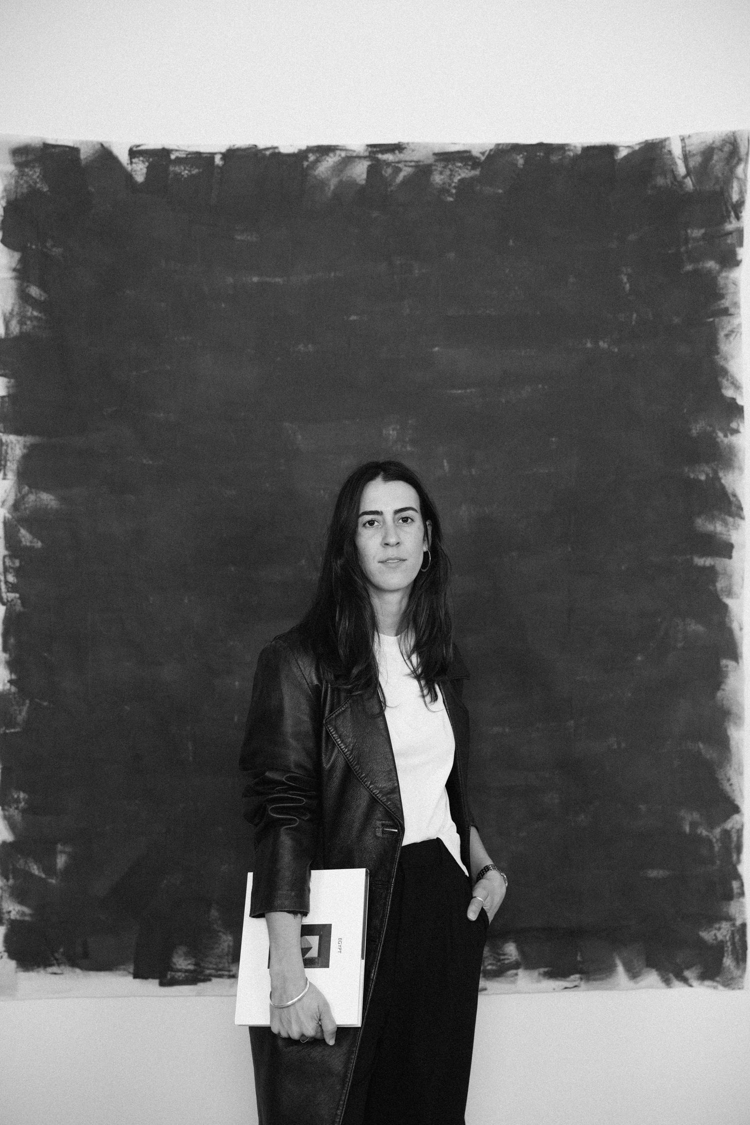 Woman in leather jacket holding a white book before a large dark painted canvas.