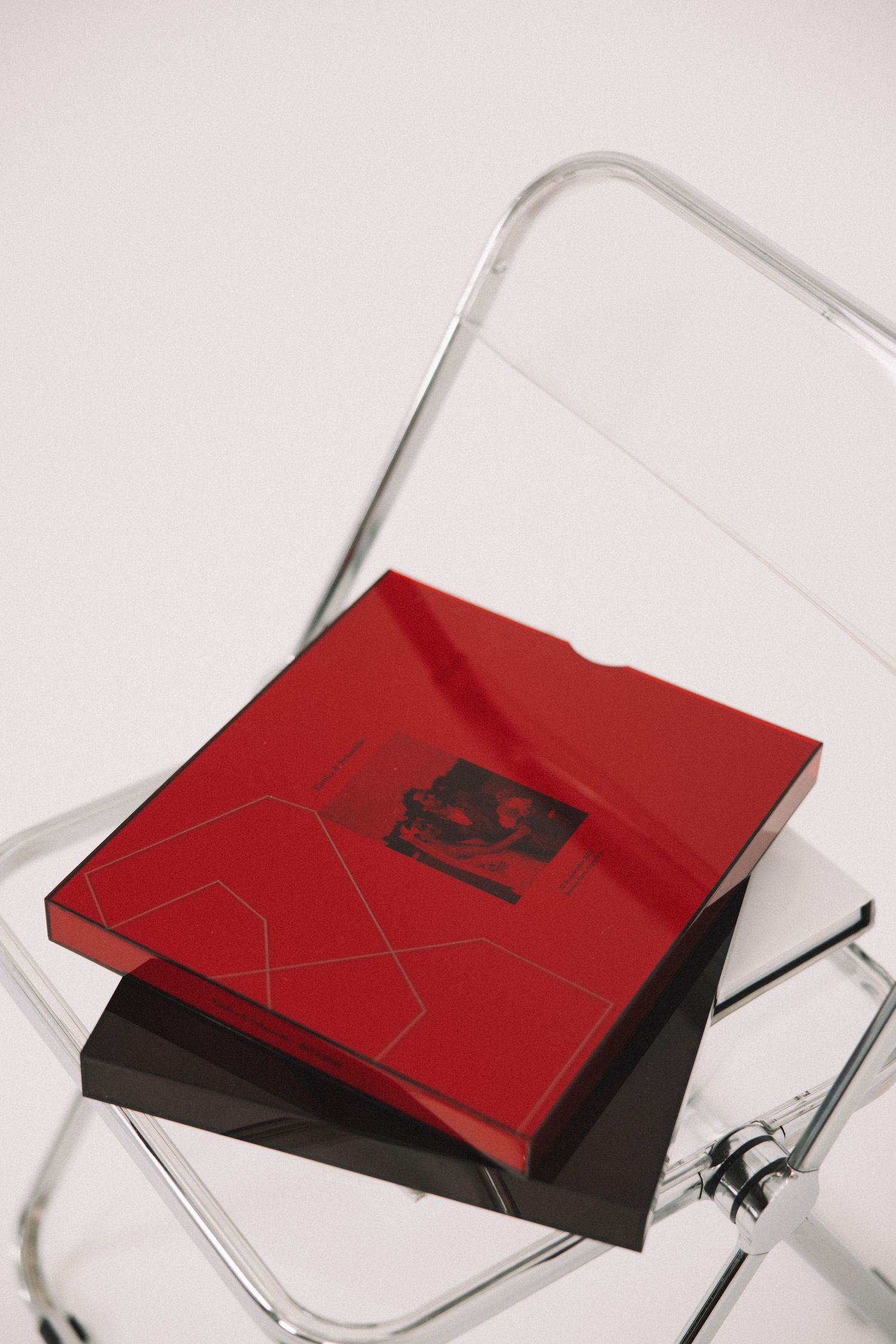 A red glossy square book sits atop a dark cover on a clear acrylic folding chair.