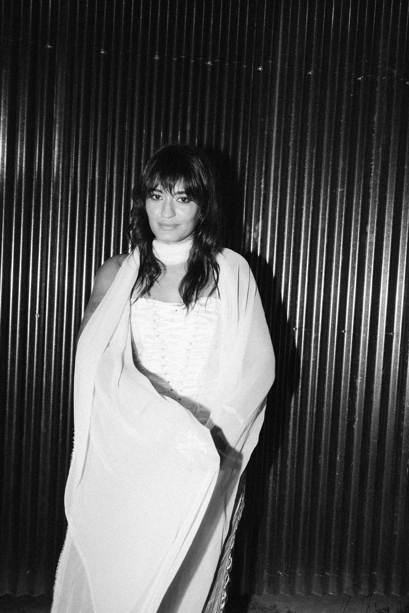 Black and white portrait of a woman in a white dress and shawl standing against a corrugated metal wall.