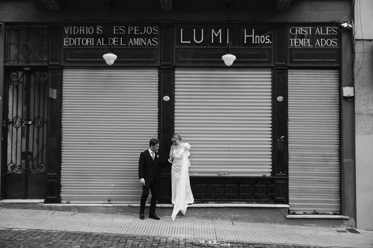 Bride and groom standing before a storefront with metal shutters on a city street.