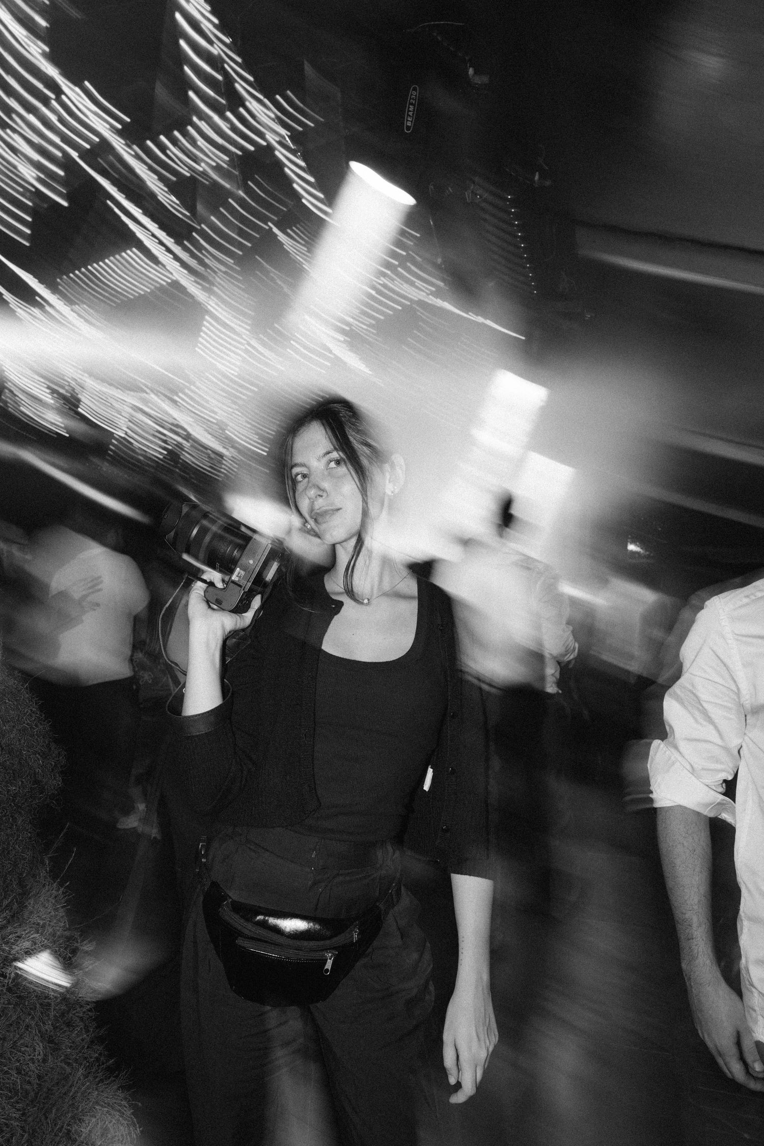 Woman holding camera in dynamic blurred nightclub scene with streaking lights in black and white.