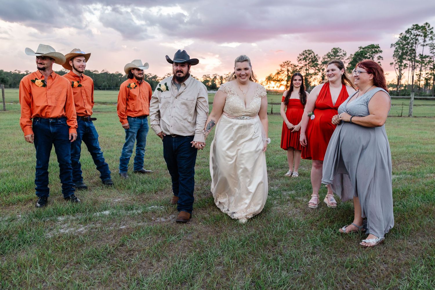 Bride and groom walk hand in hand with wedding party in orange shirts and red dresses outdoors.