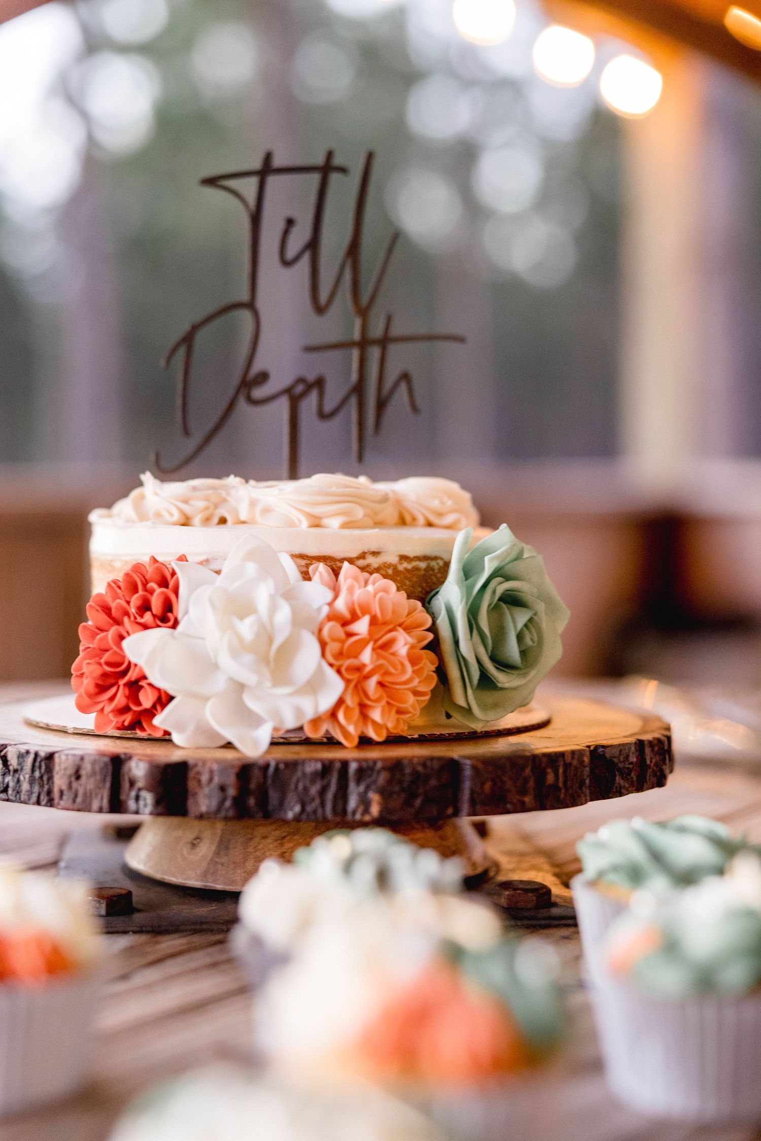 Rustic naked wedding cake topped with Till Death topper and colorful buttercream flowers on wood slice stand.