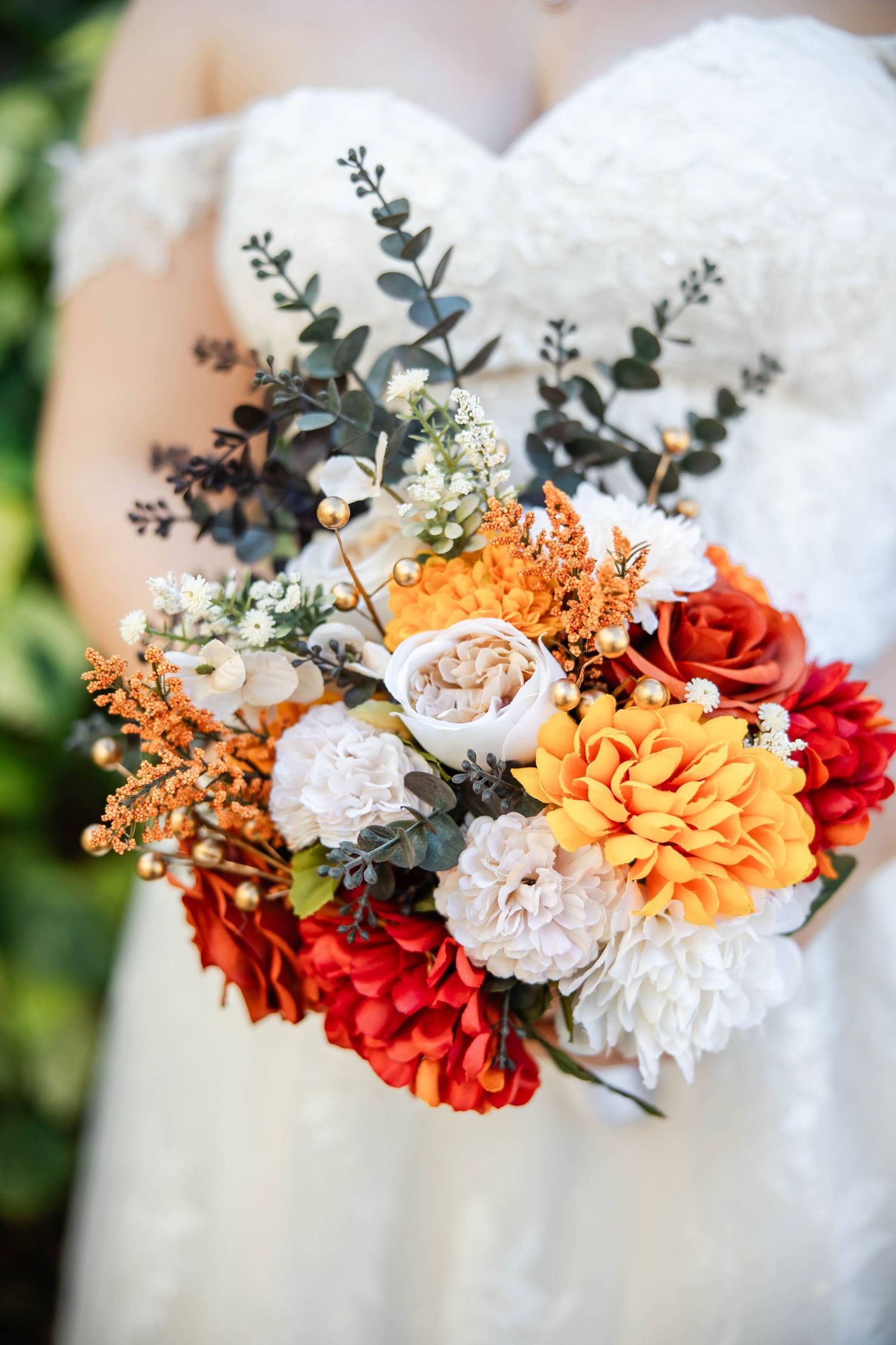 Bride holds a vibrant fall wedding bouquet with red, orange, and white flowers with greenery.