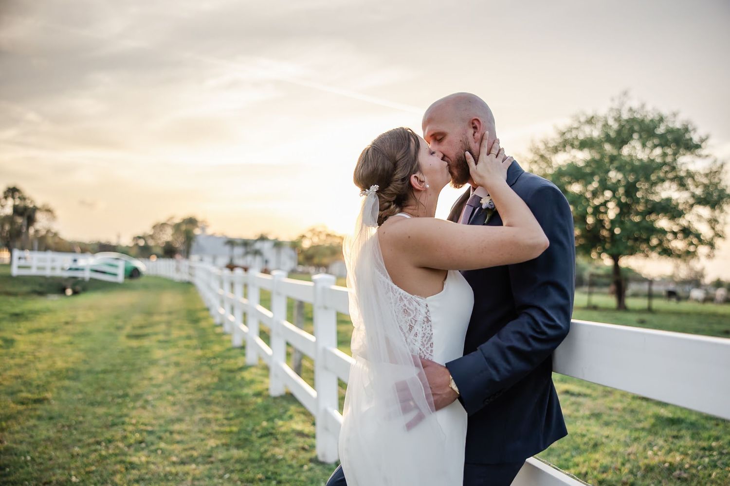 Newlyweds sharing a kiss at golden hour beside white fence at scenic outdoor wedding venue.