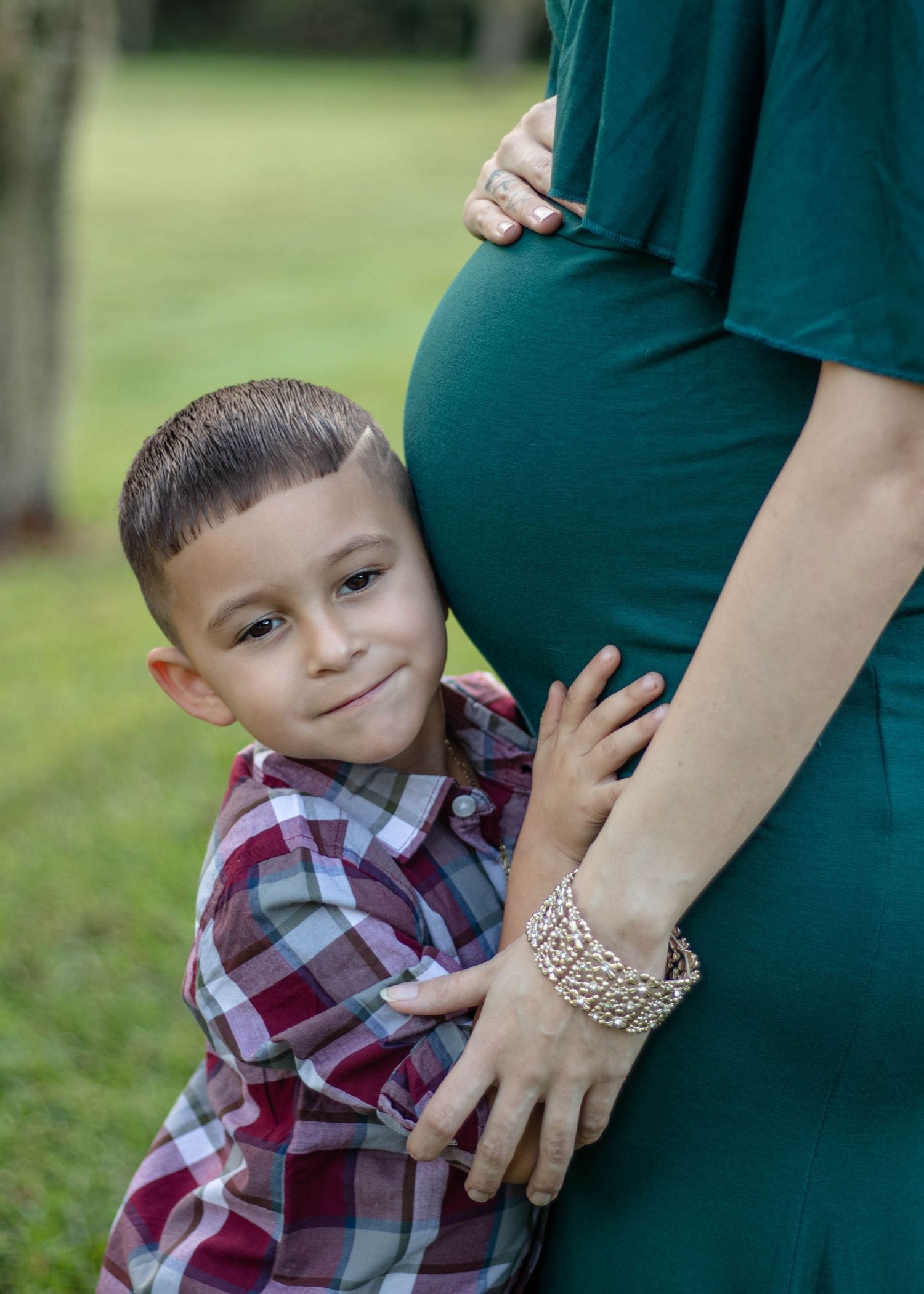 Young boy hugging pregnant mothers belly outdoors in green dress.