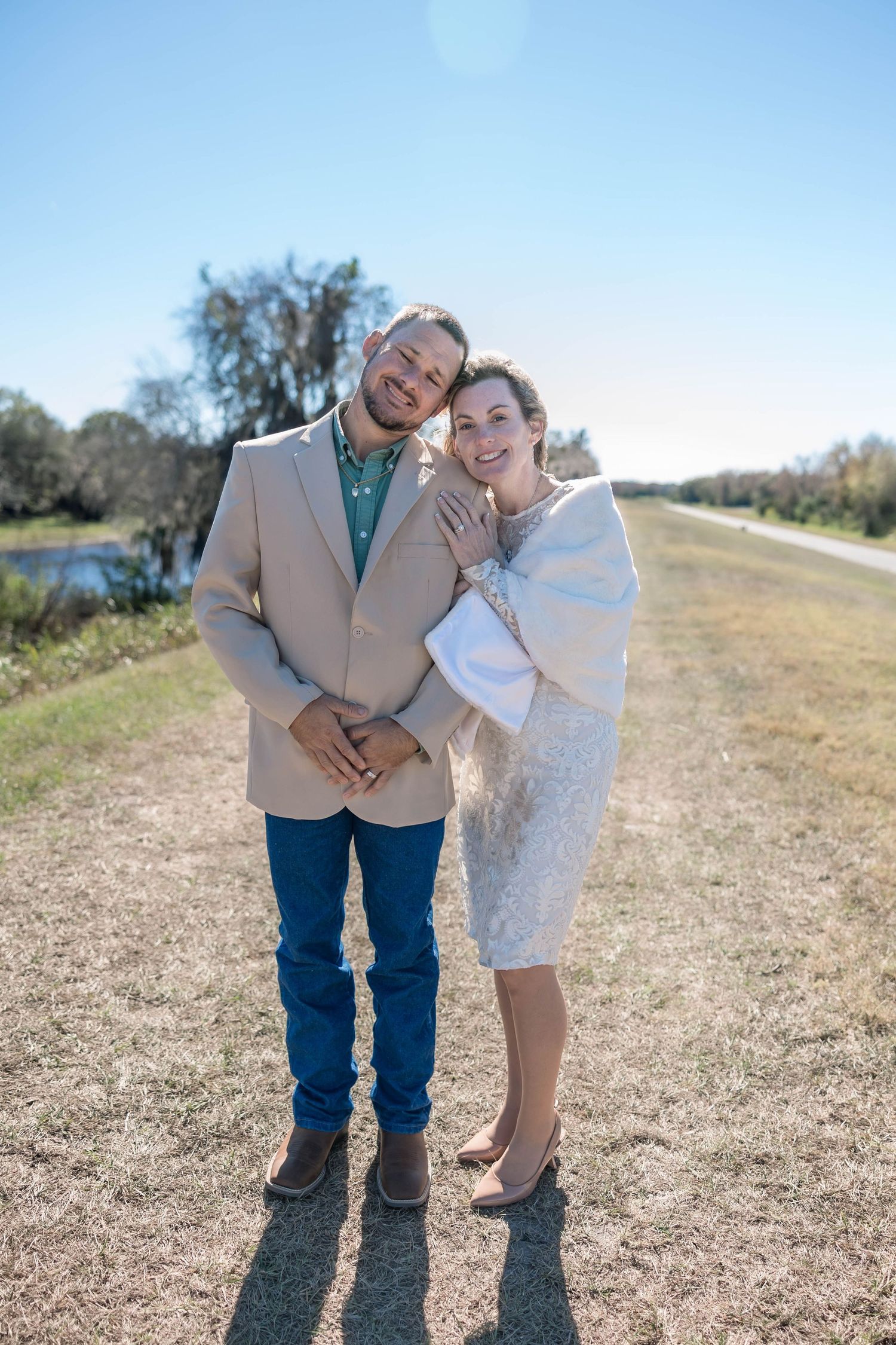 Happy couple posing together outdoors, woman in lace dress and man in beige blazer.
