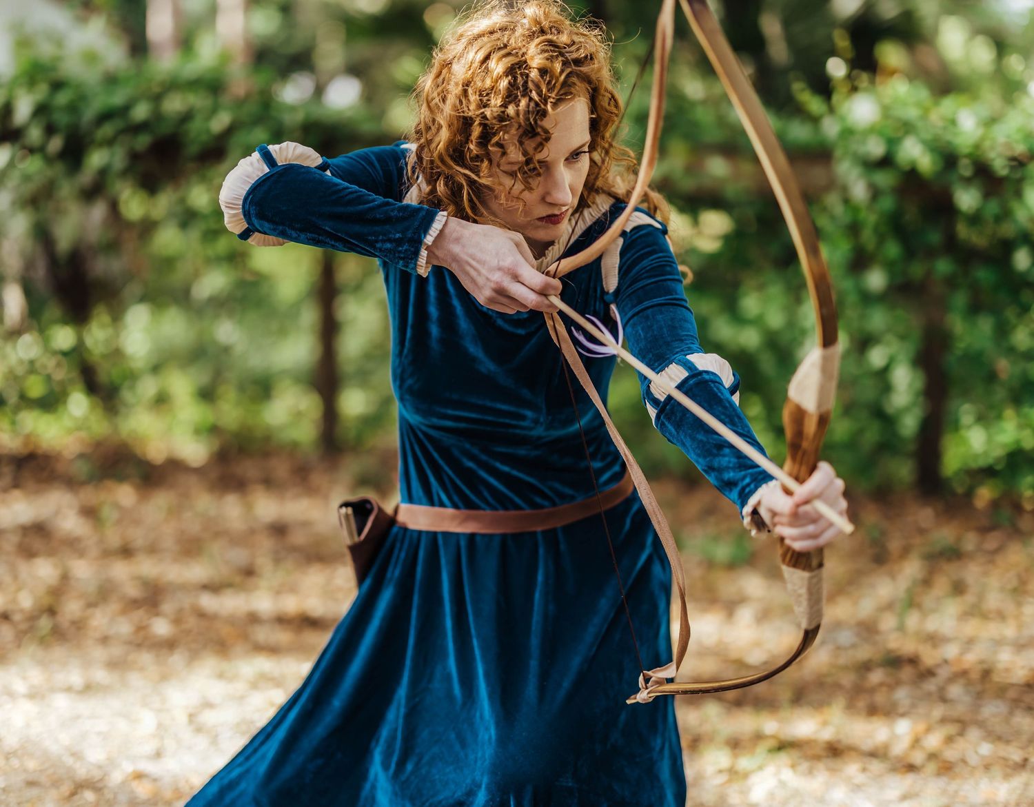 Woman in blue velvet dress drawing a bow and arrow in a forest, cosplaying as Brave's Merida.