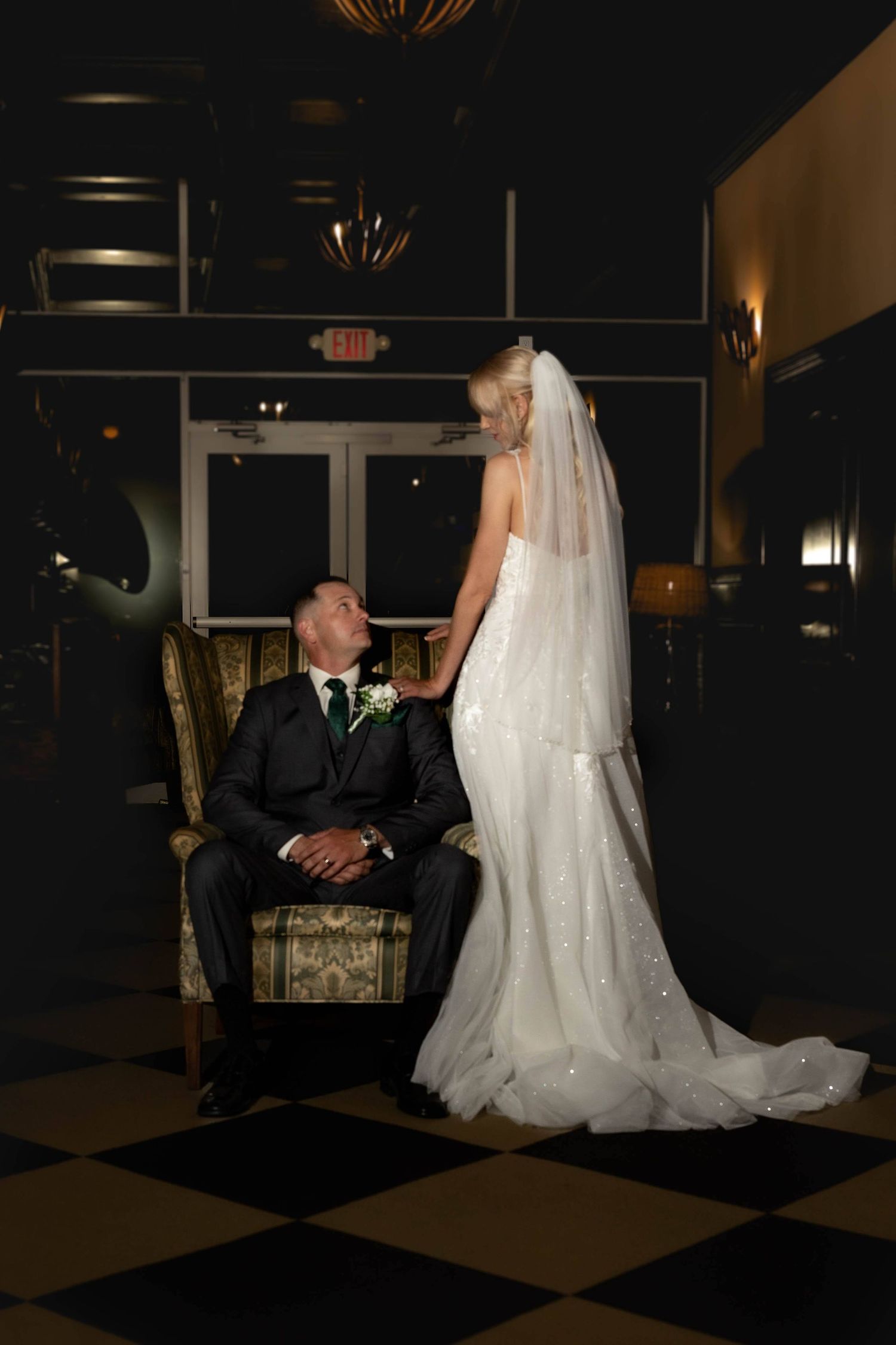 Bride in sparkle veil stands over groom seated in vintage chair in dramatic dark wedding venue.