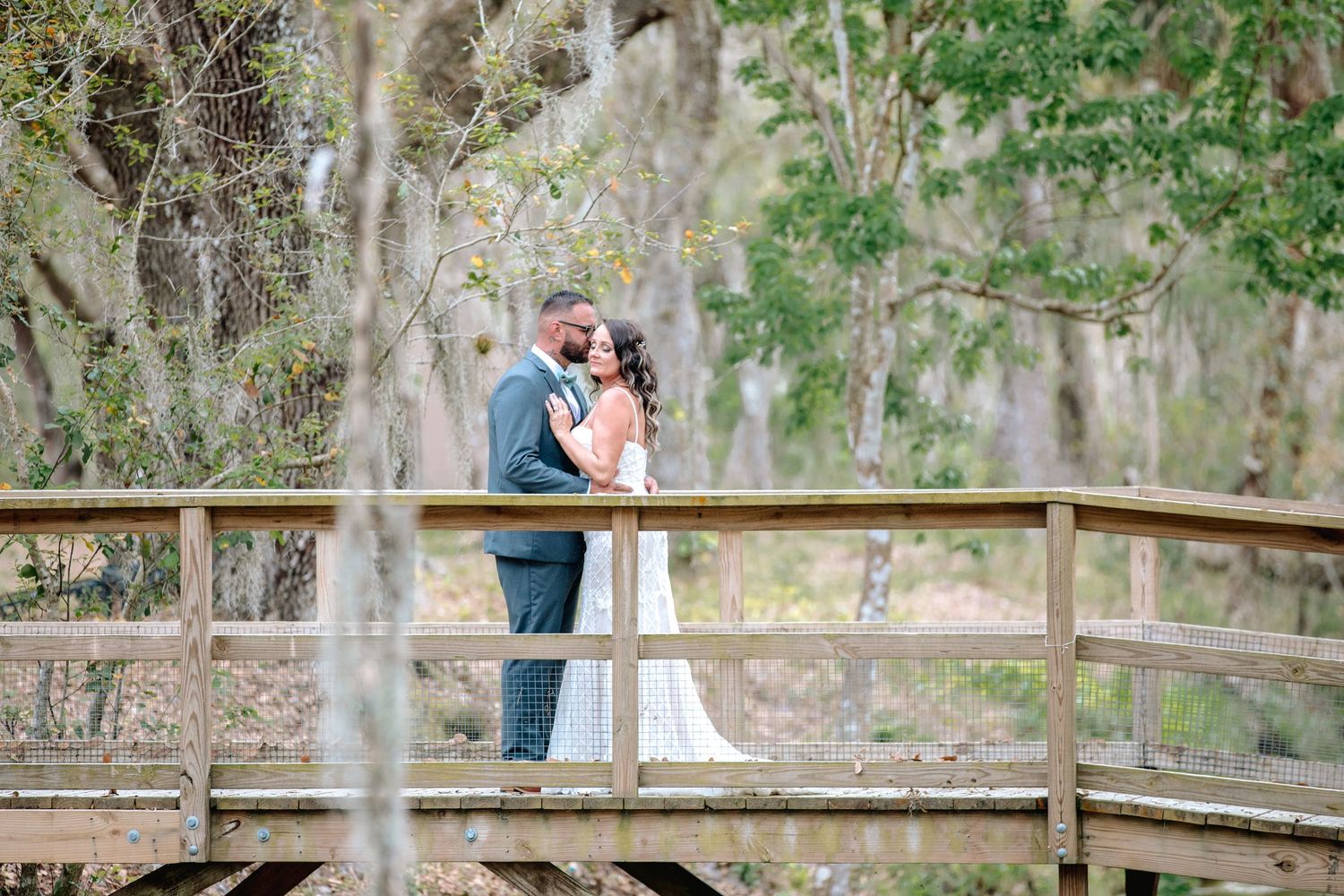 Bride and groom sharing a romantic kiss on a wooden bridge surrounded by lush trees.