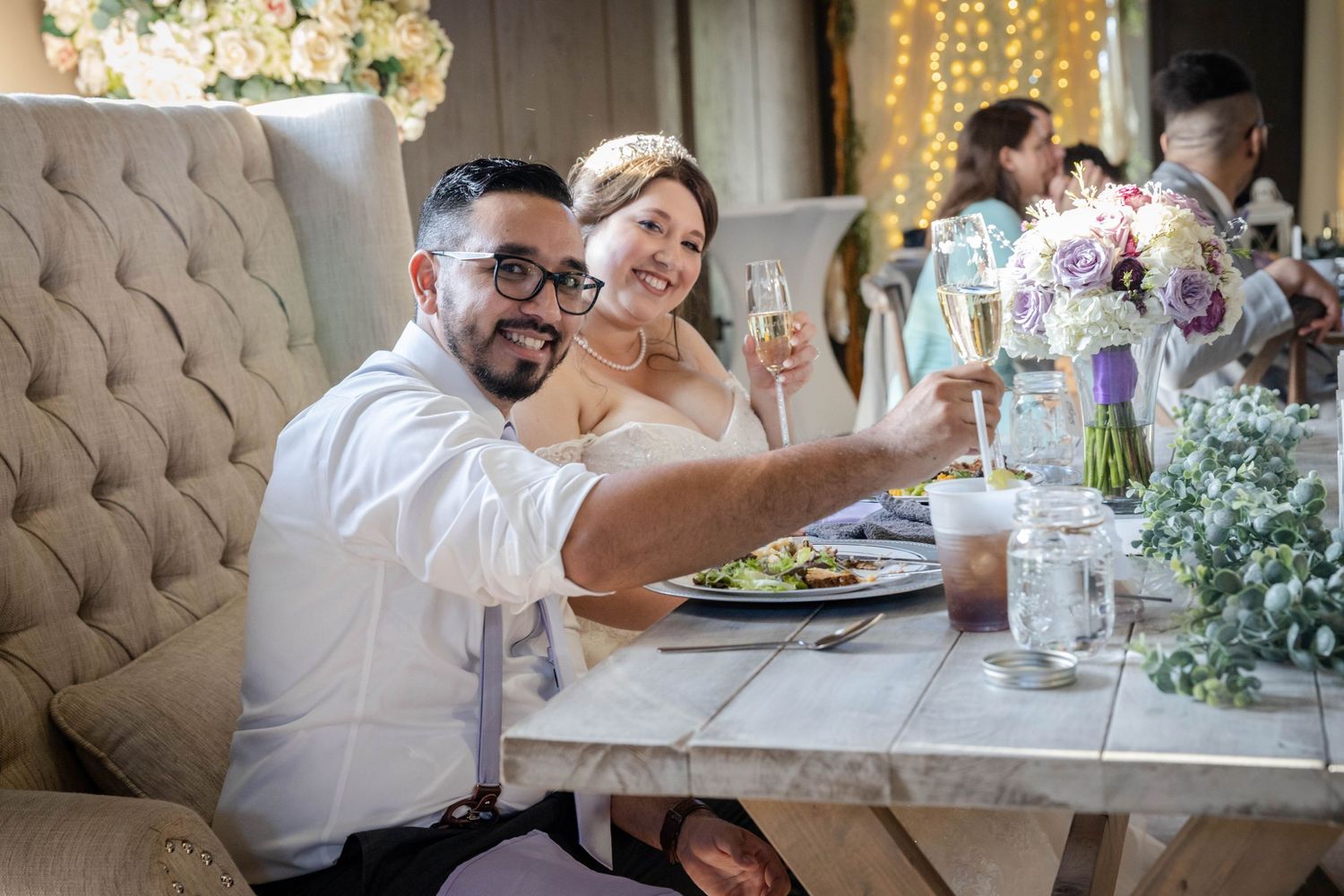 Newlywed couple smiling and raising champagne glasses at elegant floral-decorated wedding reception table.