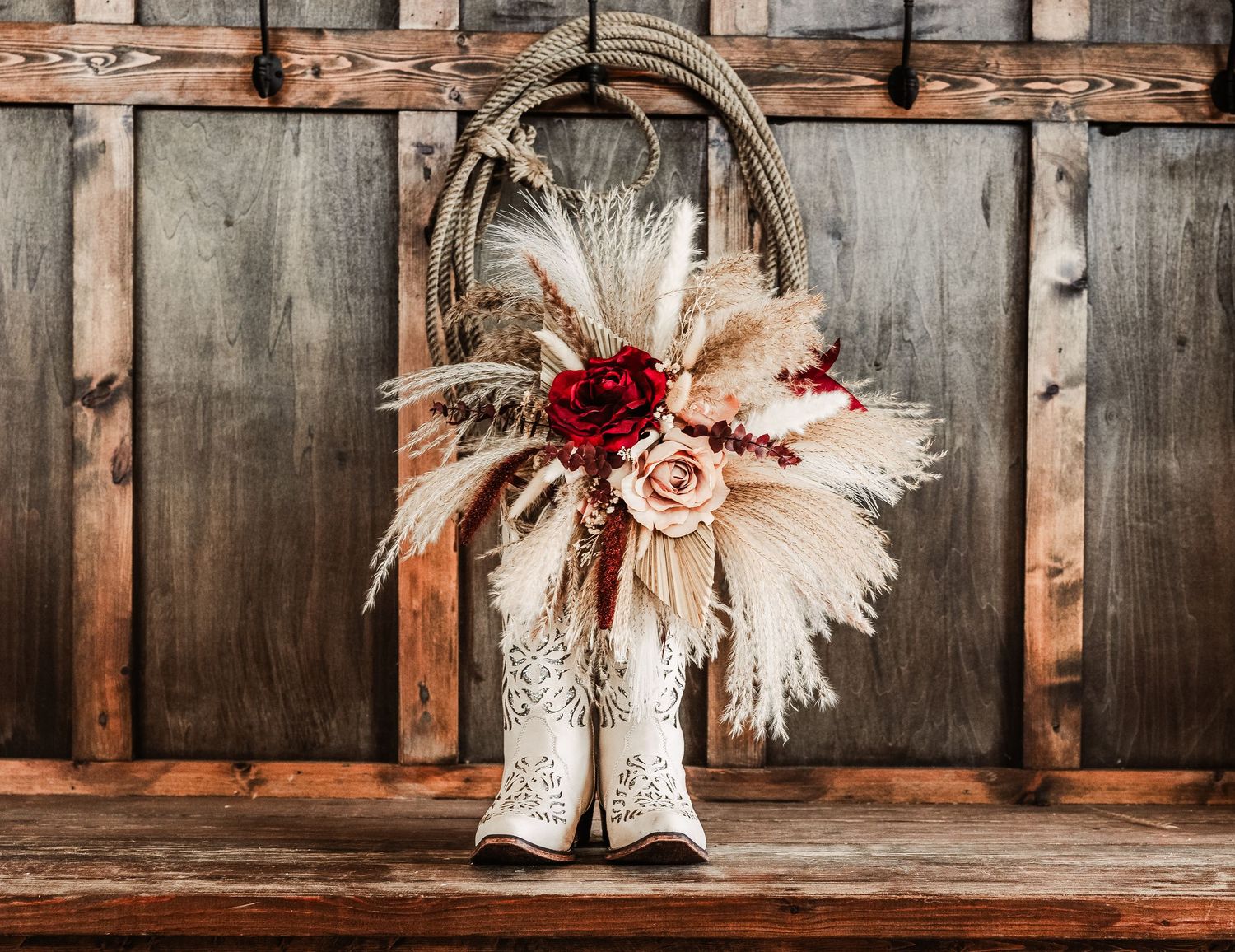 White cowboy boots with boho bouquet of pampas grass and red roses displayed on rustic wooden bench.