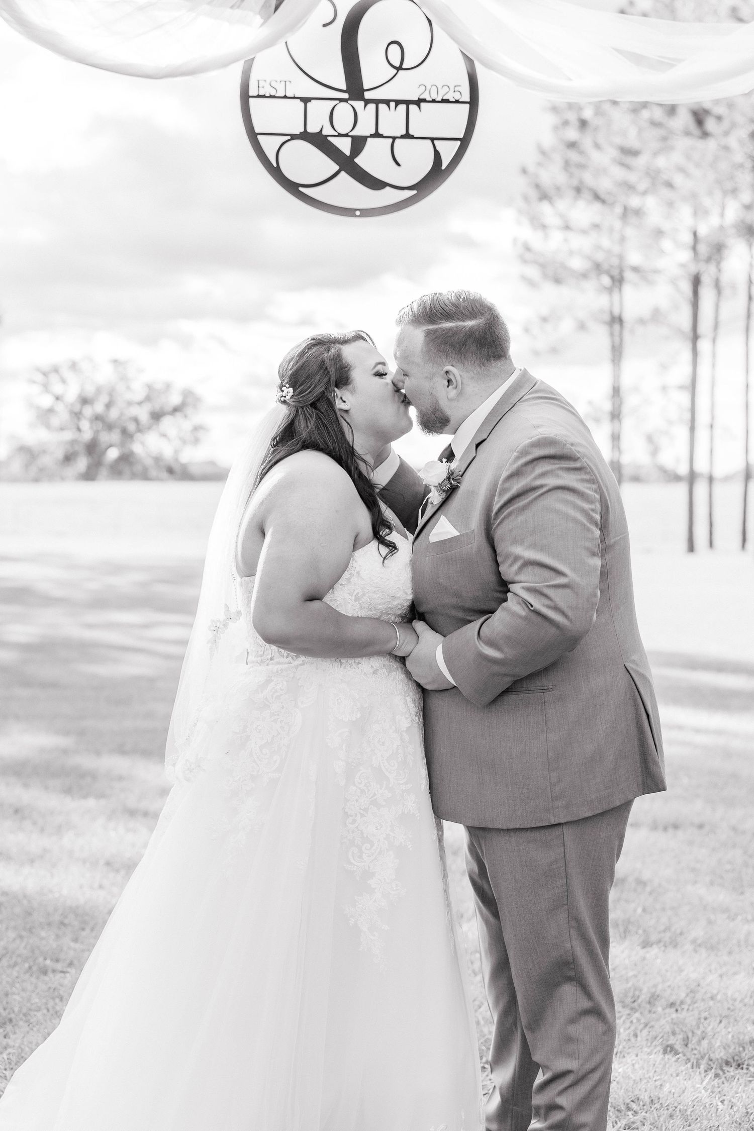 Black and white photo of bride and groom sharing first kiss under Lott family monogram sign.