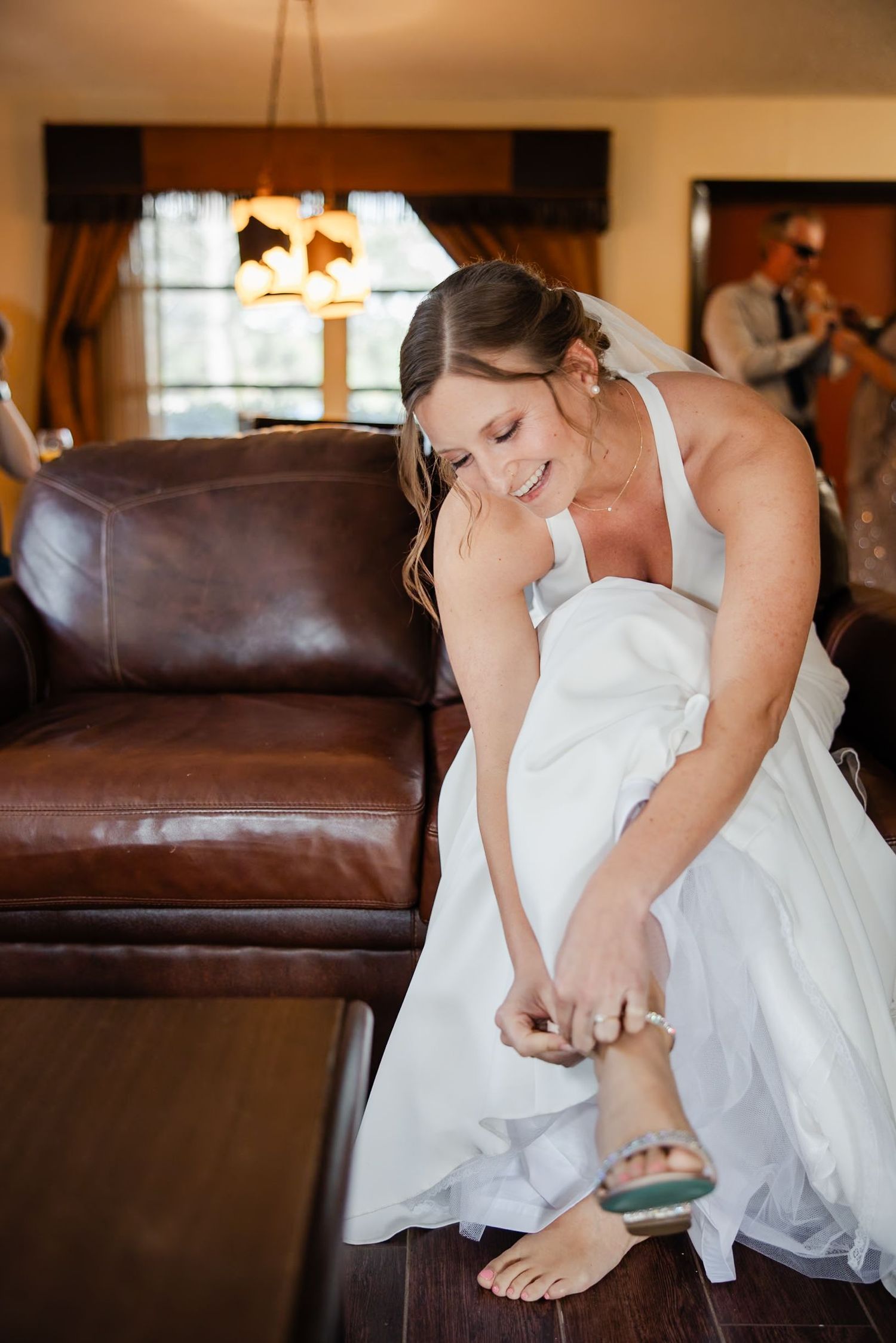 Bride in white gown smiling while putting on heeled sandal in warm rustic bridal suite.